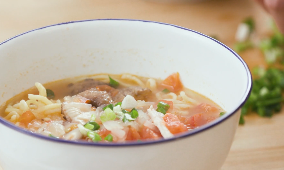 A white bowl with beef ramen soup garnished with chopped green onions and diced tomatoes.